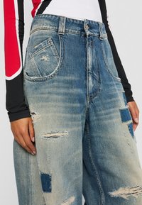 Žena v obtiahnutých modrých roztrhnutých vrecovitých džínsoch a dlhom rukáve s červenými, bielymi a čiernymi farebnými blokmi, stojí s viditeľnými rukami.