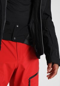 Schwarze, wetterfeste Jacke mit Reißverschluss, Druckknöpfen und verstellbaren Manschetten, kombiniert mit leuchtend roten Hosen mit Reißverschlusstaschen.