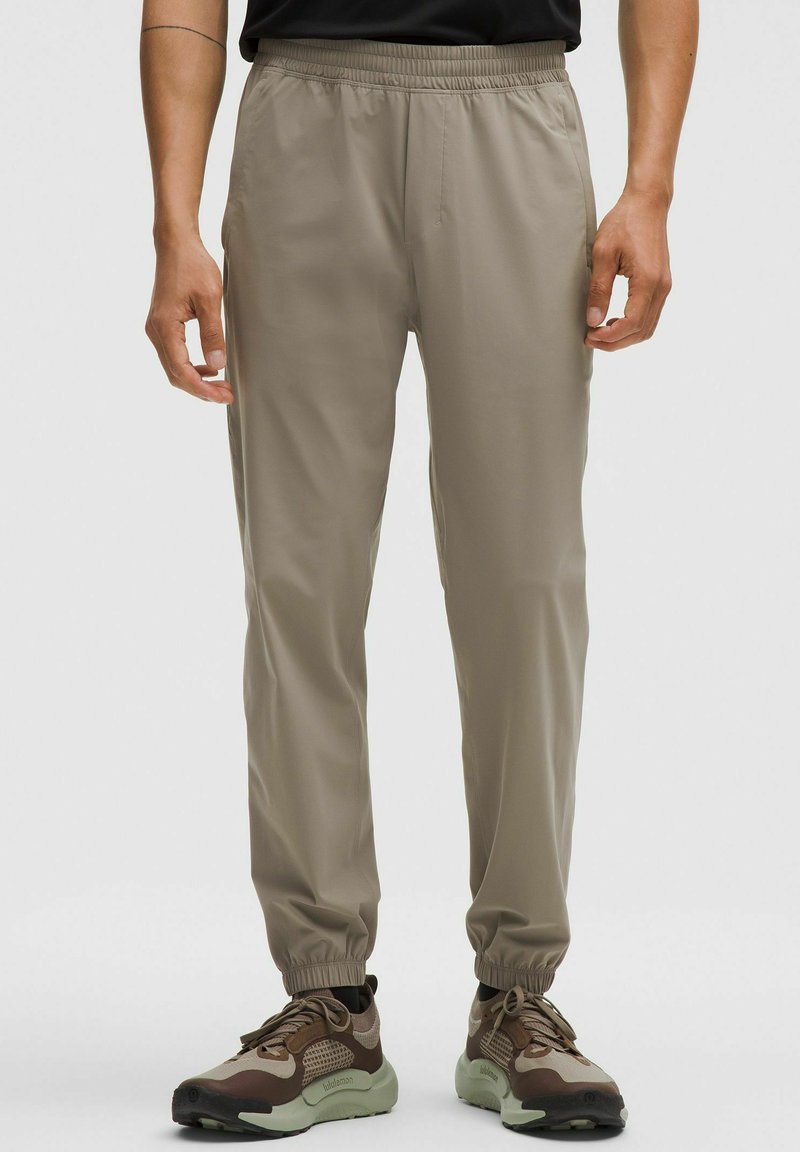 Lichte beige joggerbroeken met een elastische tailleband. Heeft een gladde textuur, enkellengte boorden en zijzakken, gecombineerd met bruine sneakers.