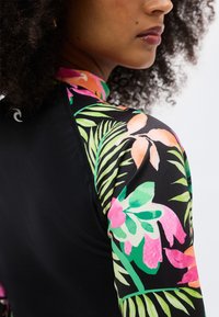 Γυναίκα με σγουρά μαλλιά που φοράει μαύρο τοπ με πολύχρωμα λουλουδένια και φυλλώδη μοτίβα στον ώμο και το μανίκι.