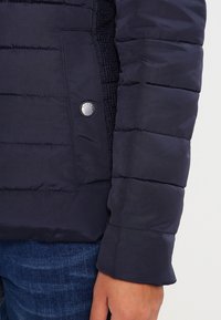 Granatowa pikowana kurtka z rękawem i kieszenią zapinaną na metalowy zatrzask, noszona z niebieskimi dżinsami, z widoczną częścią ręki.