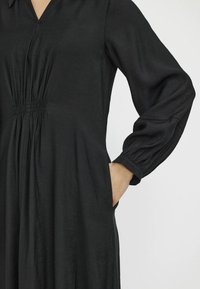 Svart klänning i mjukt tyg med V-halsringning, elastisk midja och långa voluminösa ärmar. Har sidofickor och en slät yta.