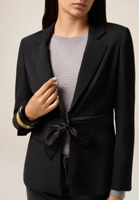 Vrouw in een zwarte blazer met een satijnen strikceintuur over een lichtgrijze geblokte trui, geaccessorieerd met twee hars armbanden om de linkerpols.