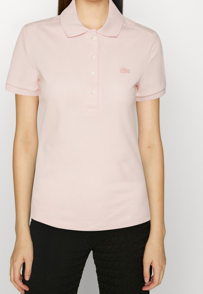 Femme portant un polo rose clair à manches courtes avec des boutons et un petit logo sur le côté gauche de la poitrine, associé à un pantalon noir.