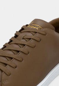 Καφέ δερμάτινο αθλητικό παπούτσι με ταίριασμα κορδονιών και λευκή σόλα, με χρυσή γραφική σήμανση στη γλώσσα.