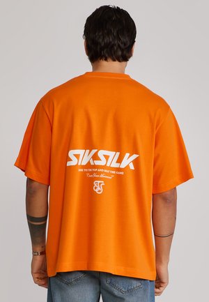 Man wearing bright orange short-sleeve shirt with white "SikSilk" logo and text on the back, paired with blue denim jeans.