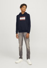 Sudadera azul marino con bolsillo frontal y logo en blanco y rojo, combinada con jeans gris claro y zapatillas blancas; un ajuste casual y materiales cómodos.