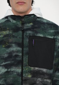 Fleece jacket with green, black, and yellow marbled patterns, featuring a zippered black chest pocket and high collar.