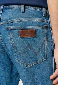 Vaqueros de mezclilla azul con un parche de cuero con un logo en relieve, costuras en contraste y un diseño de bolsillo trasero distintivo con detalle en zigzag.