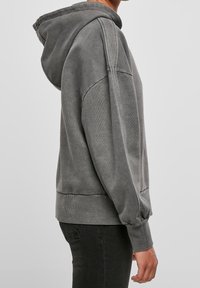 Osoba nosząca wyblakły szary hoodie z widocznym szwem na ramieniu i mankietami, w połączeniu z ciemnymi spodniami, pokazana z boku.