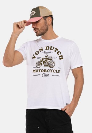 Uomo che indossa una maglietta bianca del Von Dutch Motorcycle Club e un cappellino da baseball rosa-beige, toccando la visiera del cappellino con la mano sinistra.