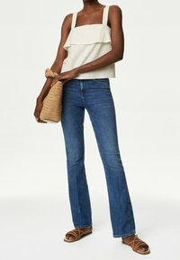 Cremfarbener Top mit quadratischem Ausschnitt und geschichteten Volants, kombiniert mit dunkelblauen Schlagjeans und braunen Riemensandalen. Hält eine geflochtene Handtasche.