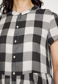 Robe à carreaux noir et blanc en tissu léger, avec un col rond, des manches courtes et des boutons décoratifs sur le devant.