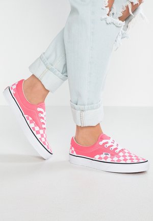 Pieds en jean bleu clair retroussé et déchiré, portant des baskets Vans à carreaux roses et blancs, sur un sol blanc.