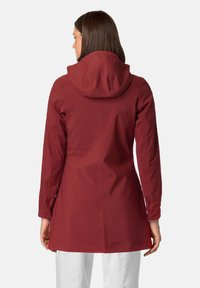 Vrouw in een maroon, knielange, langmouwige hoodie gezien vanuit de achterkant, gecombineerd met witte broek tegen een effen achtergrond.