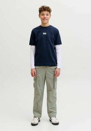 Teismeline lokkis juustega, kandes tumesinist lühikeste varrukatega särki valgete pika varrukaga all, heledaid cargo-pükse ja tossusid, seisab ühtlasel taustal.