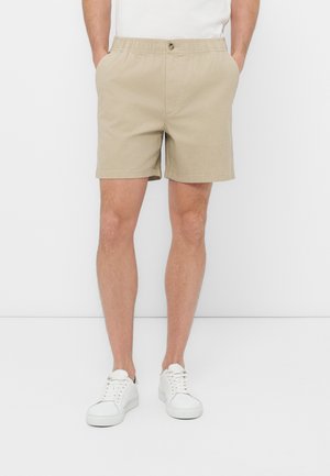 Muž nosící béžové kraťasy s elastickým pasem, bílé tenisky a bílé tričko, stojící s rukama v kapsách na bílém pozadí.