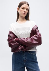 Νεαρή γυναίκα με ίσια καστανά μαλλιά σταυρώνει τα χέρια της, φοράει λευκό t-shirt DKNY και εν μέρει κατεβασμένο μπουφάν από δέρμα σε μπορντό χρώμα, συνδυασμένο με μπλε τζιν.