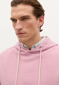 Lichtroze hoodie van zacht materiaal, met een trekkoord capuchon, gecombineerd met een gestreepte blouse met kraag eronder. Subtiel merkdetail.