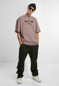 Oversized beige t-shirt with printed logo, paired with black cargo pants featuring pockets. White sneakers and a black beanie complete the look.
