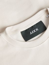 Felpa beige con colletto rotondo. Il tessuto ha una texture morbida. Etichetta nera con la scritta "JUXX" stampata in bianco, etichetta della taglia visibile.