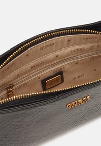 Sort struktureret lædertaske indeni, prydet med en beige foring med "GUESS" tryk og en gylden lynlås. Indeholder en lynlåssektion og et logomærke.