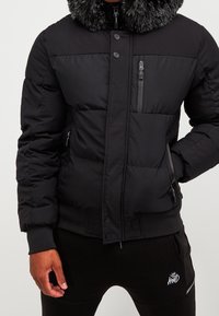 Černá péřová bunda s límcem z umělé kožešiny, zipovou kapsou na hrudi a žebrovanými manžetami. Hladký materiál a detail s knoflíkovým uzavřením.