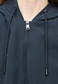 Κοντινό πλάνο φερμουάρ σκούρου μπλε μπουφάν με κουκούλα, μεταλλική λαβή και δύο κορδόνια με μεταλλικές άκρες, που φοριέται στο λαιμό και το πάνω μέρος του στήθους ενός ατόμου.