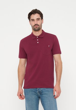 Mann trägt ein burgunderfarbenes, kurzärmeliges Polohemd mit Knöpfen und einem kleinen Taschendetail, kombiniert mit blauen Jeans, steht vor weißem Hintergrund.