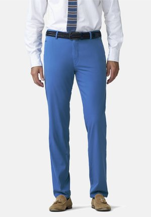 Man draagt een blauwe broek, wit overhemd, gestreepte stropdas, zwarte gevlochten riem en beige suède loafers met kwastjes, staand in een rechte houding.