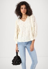 Blusa in pizzo color avorio con scollatura a V, maniche a sbuffo e motivo floreale. Abbinata a jeans attillati blu chiaro e una borsa nera a fiori.