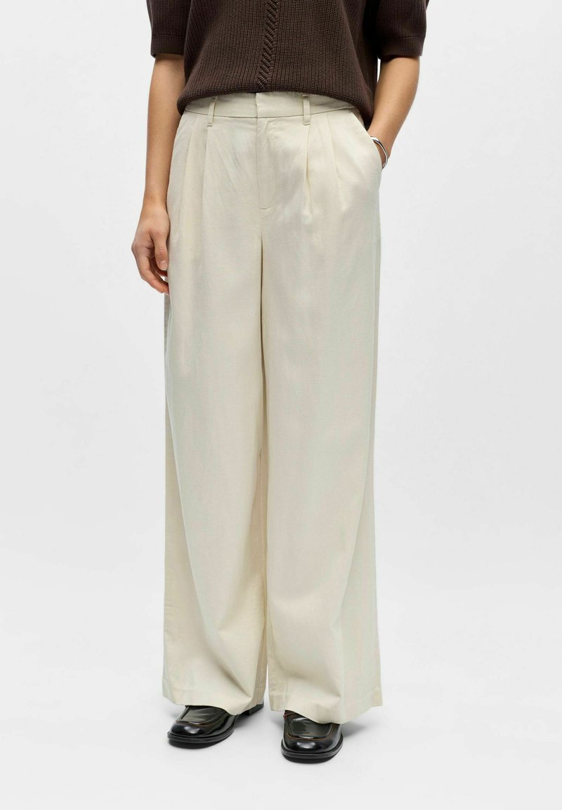 Femme portant un pantalon crème taille haute à jambes larges, des mocassins noirs et un haut en tricot manches courtes marron foncé, debout les mains dans les poches.