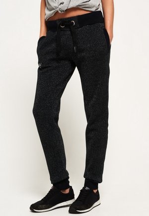 Person trägt dunkelgraue gesprenkelte Jogginghose mit schwarzem Bund und Bündchen, Hände in den Taschen, kombiniert mit schwarzen Sneakers vor hellem Hintergrund.