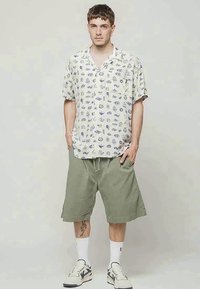 Camisa de mangas curtas em creme claro com ilustrações em azul escuro; combinada com calções verdes-oliva, meias brancas e ténis.