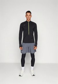 Fekete, sárga cipzárral ellátott sportos cipzáras dzseki, szürke rövidnadrág és fekete leggings. Fehér sportcipő teszi teljessé a sportos öltözéket, semleges háttér előtt.