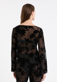 Woman with long brown hair wearing a sheer black long-sleeve top and matching pants with velvet floral patterns, viewed from the back.