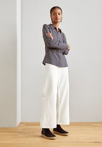 Gemustertes schwarz-weißes Hemd mit langen Ärmeln, kombiniert mit weißen, weiten Hosen und dunklen Knöchelstiefeln.