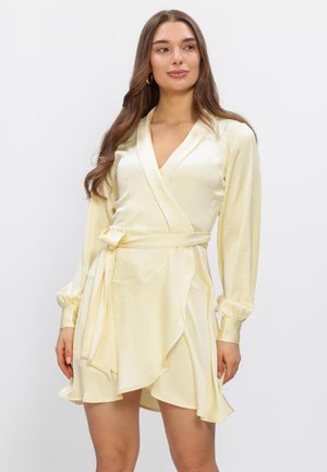 Vrouw met lang bruin haar, gekleed in een lichtgele satijnen wikkeljurk met lange mouwen en een gestrikte ceintuur, staand tegen een effen achtergrond.
