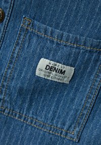 Tessuto in denim blu con etichetta bianca. L'etichetta riporta la scritta "NAME IT DENIM ORIGINAL QUALITY SINCE 1996." Presenta cuciture e dettagli della tasca.