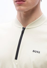 Κοντινό πλάνο ενός ατόμου που φοράει ένα ανοιχτό μπεζ πουκάμισο  με φερμουάρ και μαύρο φερμουάρ, με το λογότυπο "BOSS" στο στήθος.