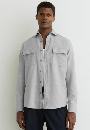 Homme portant une chemise gris clair à boutons sur un haut blanc et un pantalon foncé, debout devant un fond blanc uni.