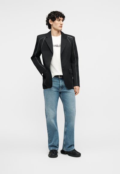 Άνδρας με σγουρά μαλλιά, φοράει μαύρο σακάκι με λευκές ραφές, λευκό μπλουζάκι με γραφικό σχέδιο, μπλε τζιν και μαύρα αθλητικά παπούτσια, στέκεται με το ένα χέρι στην τσέπη.