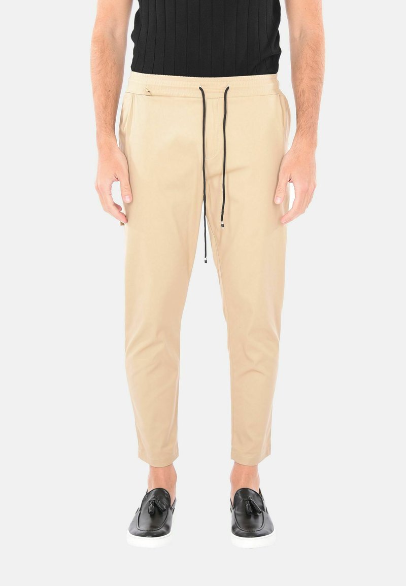 Pantaloni beige a sigaretta in tessuto leggero con vita elastica, coulisse nera e due tasche laterali, abbinati a scarpe nere senza stringhe.