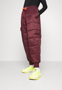 Burgunderfarbene gepolsterte Hose mit einem elastischen Bund, seitlichen Taschen und roten Akzenten, kombiniert mit klobigen neon-gelben Sneakers.