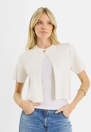 Mujer rubia con un cárdigan blanco de manga corta con un solo botón sobre una camiseta blanca y jeans azules, con las manos en los bolsillos, mirando ligeramente hacia la derecha.