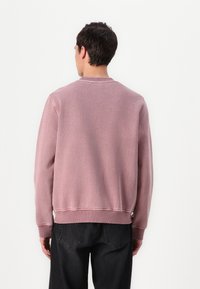 Rosa Sweatshirt mit Rundhalsausschnitt, strukturiertem Stoff und gerippten Bündchen. Getragen mit schwarzen Denim-Hosen, die eine entspannte Passform und ein minimalistisches Design zeigen.