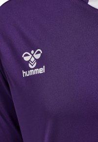 Fioletowa tkanina sportowego jersey z białym logo hummel przedstawiającym stylizowaną pszczołę i tekst na obszarze klatki piersiowej.
