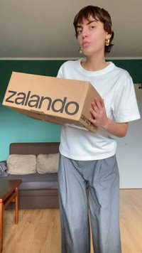 Kartoninė dėžė su juodu užrašu "zalando", laikoma prieš žmogų, dėvintį baltą marškinėlį ir pilkas plačias kelnes, su sofa fone.