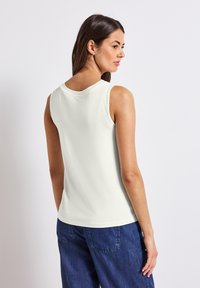 Top bianco senza maniche con scollatura rotonda, tessuto liscio, indossato con pantaloni in denim blu. La vista posteriore mostra linee pulite e un design semplice.