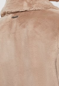 Mjuk beige fuskpälsjacka med vikbar krage och en liten rektangulär metallbrandtag nära axeln.